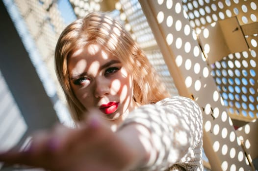 Portrait of a young woman dressed in a knitted sweater and blue jeans. Standing near a metal perforated parking fence construction. Shadows in the form of dots of light fall on her face