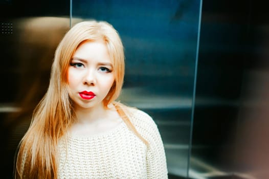 Portrait of a female face, young blond woman with plump lips and beautiful eyes dressed in a knitted sweater. Against elevator lift background