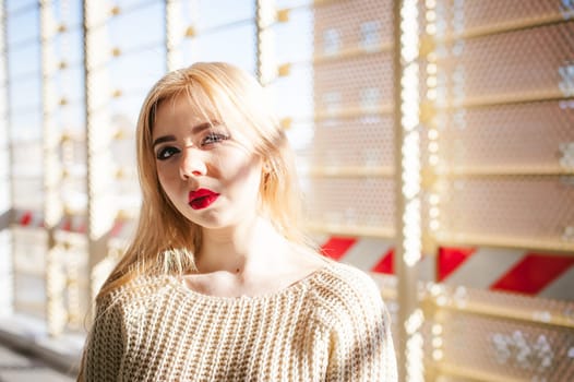 Portrait of a young woman dressed in a knitted sweater and blue jeans. Standing near a metal perforated parking fence construction