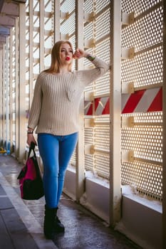 Portrait of a young woman dressed in a knitted sweater and blue jeans, holds in her hand a sports bag. Standing near a metal perforated parking fence construction
