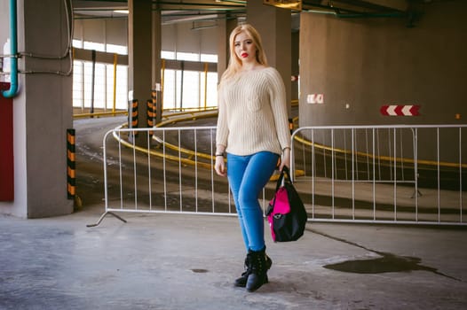Portrait of a young woman dressed in a knitted sweater and blue jeans, holds in her hand a sports bag. standing on the background of a climb to the parking floor