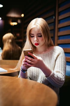 Young blond woman in a knitted sweater, sitting at a table in a cafe, spending time alone, doing internet communication through wifi