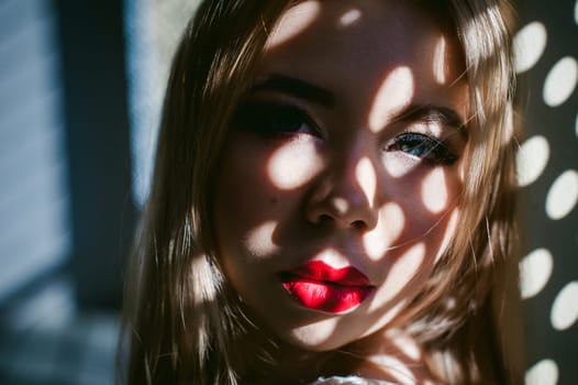 Portrait of a young woman dressed in a knitted sweater and blue jeans. Standing near a metal perforated parking fence construction. Shadows in the form of dots of light fall on her face