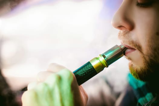 young vaper man with beard vaping mechanical mod. Guy smokes an electronic cigarette by blowing a smoke vapor. Bringing rda mouthpiece to the lips on colorful background