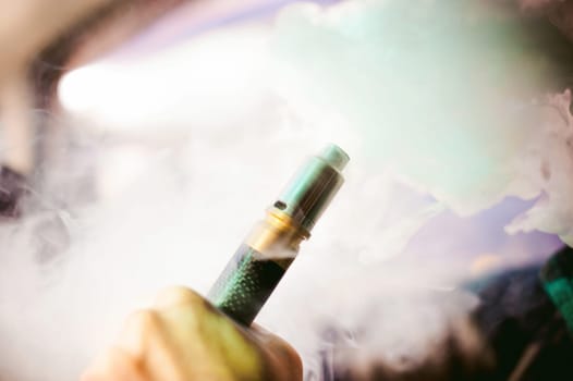 young vaper man with beard vaping mechanical mod. Guy smokes an electronic cigarette by blowing a smoke vapor. Holds in hand on colorful background