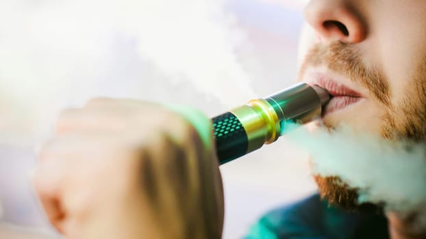 young vaper man with beard vaping mechanical mod. Guy smokes an electronic cigarette by blowing a smoke vapor. Bringing rda mouthpiece to the lips on colorful background