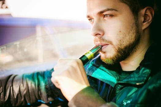 young vaper man with beard vaping mechanical mod. Guy smokes an electronic cigarette by blowing a smoke vapor. Bringing rda mouthpiece to the lips on colorful background