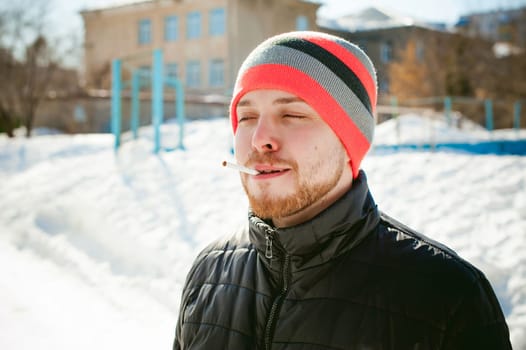 Portrait of a man with a beard with a mustache, smoking a cigarette, with an emotional face in the open air. Dressed in a bright striped hat, against the background of a snowy sunny day