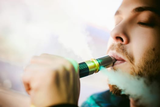young vaper man with beard vaping mechanical mod. Guy smokes an electronic cigarette by blowing a smoke vapor. Bringing rda mouthpiece to the lips on colorful background