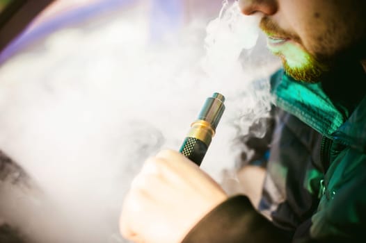 young vaper man with beard vaping mechanical mod. Guy smokes an electronic cigarette by blowing a smoke vapor. Holds in hand on colorful background