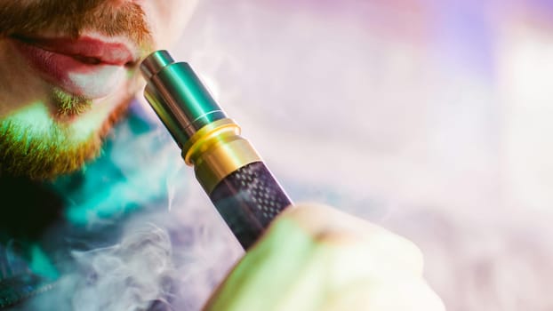 young vaper man with beard vaping mechanical mod. Guy smokes an electronic cigarette by blowing a smoke vapor. Bringing rda mouthpiece to the lips on colorful background