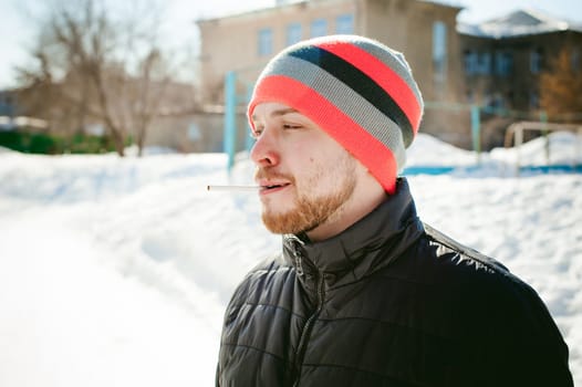 Portrait of a man with a beard with a mustache, smoking a cigarette, with an emotional face in the open air. Dressed in a bright striped hat, against the background of a snowy sunny day