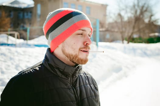 Portrait of a man with a beard with a mustache, smoking a cigarette, with an emotional face in the open air. Dressed in a bright striped hat, against the background of a snowy sunny day