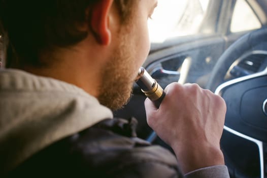 young vaper man with beard vaping mechanical mod. Guy smokes an electronic cigarette by blowing a smoke vapor. Bringing rda mouthpiece to the lips on colorful background Sitting at the wheel of a car