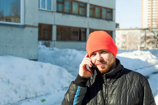 man Emotionally communicates by telephone. guy with beard, dressed in an autumn sports casual jacket and bright orange hat, Speaks by cell phone in stylish case, walking outdoors on sunny winter day