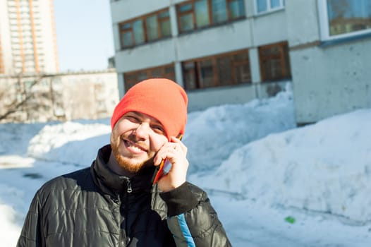 man Emotionally communicates by telephone. guy with beard, dressed in an autumn sports casual jacket and bright orange hat, Speaks by cell phone in stylish case, walking outdoors on sunny winter day