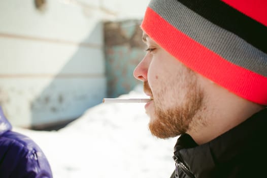 Portrait of a man with a beard with a mustache, smoking a cigarette, with an emotional face in the open air. Dressed in a bright striped hat, against the background of a snowy sunny day