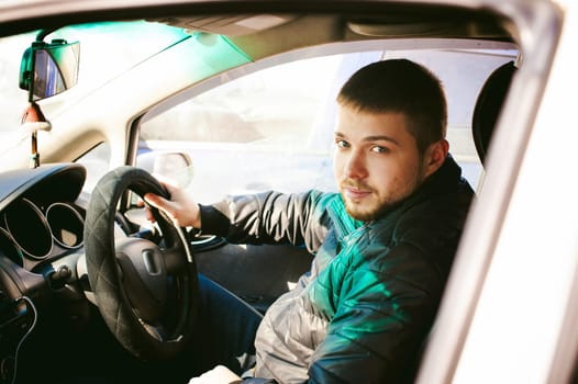 young male driver sits holding his hand behind the wheel in the salon of his car, a happy look of a bearded motorist man