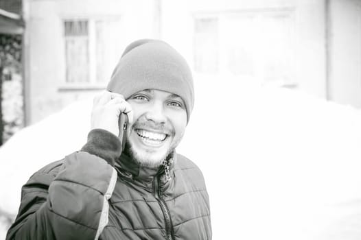 Telephone calls men. The guy is talking on cell phone in street in winter, dressed in warm clothes, holds in his hand phone at ear, emotionally smiling