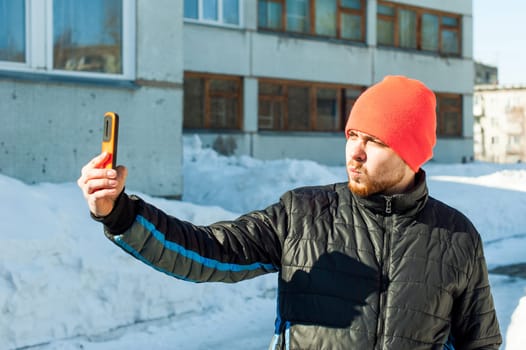 Man photographing himself. guy with beard, dressed in an autumn sports casual jacket and bright orange hat, makes selfie on phone in stylish case, walking outdoors on sunny winter day