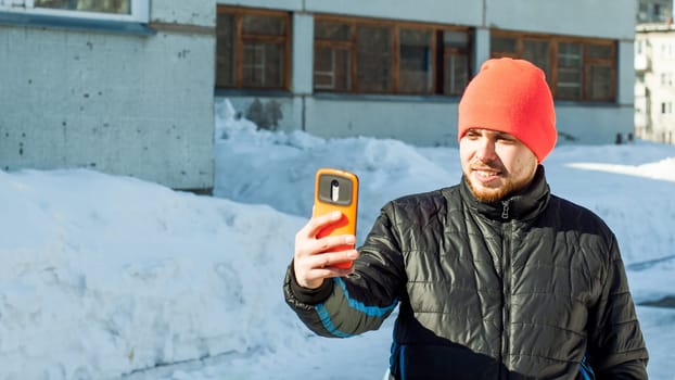 Man photographing himself. guy with beard, dressed in an autumn sports casual jacket and bright orange hat, makes selfie on phone in stylish case, walking outdoors on sunny winter day