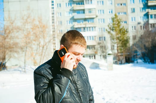 Telephone calls men. The guy is talking on cell phone in street in winter, dressed in warm clothes, holds in his hand phone at ear, in bright orange sheath case