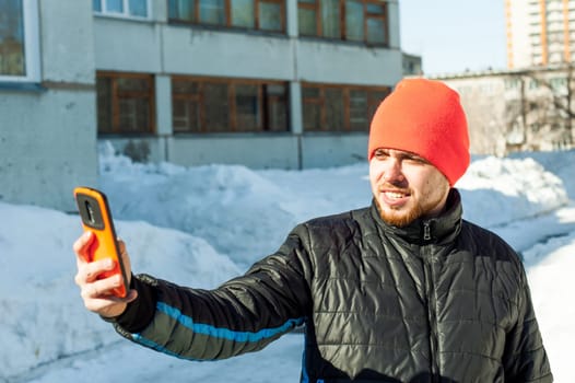 Man photographing himself. guy with beard, dressed in an autumn sports casual jacket and bright orange hat, makes selfie on phone in stylish case, walking outdoors on sunny winter day