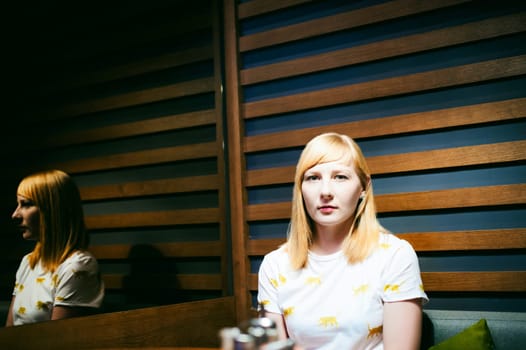 Young blonde woman wearing white T-shirt with print, visited cafe lunch time, drinking fruity hot drink from glass teapot white cup hand holding, background stylized tree table lit up with warm light