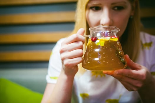 Young blonde woman wearing white T-shirt with print, girl Holds a glass jug in his hand, sitting at a table in a cafe, background stylized Wood texture lit up with warm light