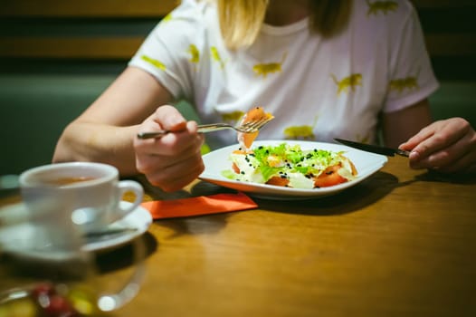 Young blonde woman wearing white T-shirt with print, girl Eats salad at lunch time, sitting at a table in a cafe, background stylized Wood texture lit up with warm light