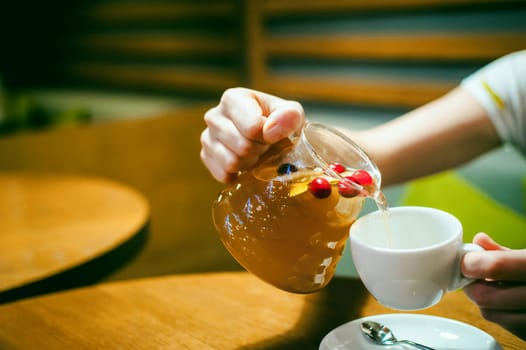 Young blonde woman wearing white T-shirt with print, girl pours a fruit drink from a glass jug, into a white cup, sitting at a table in a cafe, background stylized Wood texture lit up with warm light