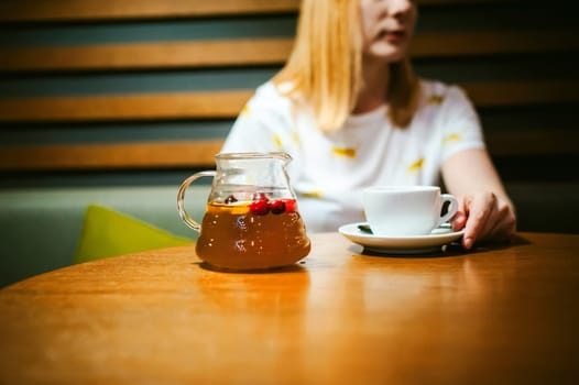 Young blonde woman wearing white T-shirt with print, visited cafe lunch time, drinking fruity hot drink from glass teapot white cup hand holding, background stylized tree table lit up with warm light
