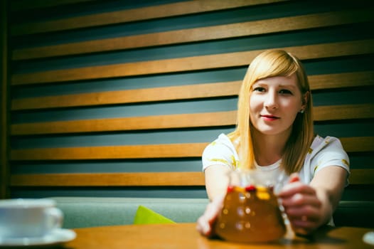 Young blonde woman wearing white T-shirt with print, girl Holds a glass jug in his hand, sitting at a table in a cafe, background stylized Wood texture lit up with warm light