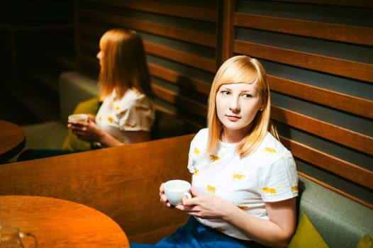 Young blonde woman wearing white T-shirt with print, visited cafe lunch time, drinking fruity hot drink from glass teapot white cup hand holding, background stylized tree table lit up with warm light