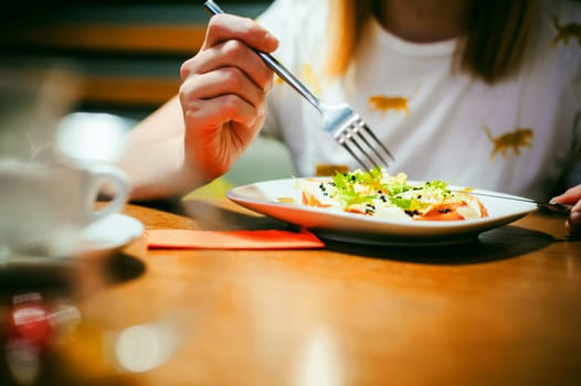 Young blonde woman wearing white T-shirt with print, girl Eats salad at lunch time, sitting at a table in a cafe, background stylized Wood texture lit up with warm light