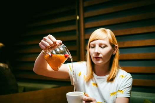 Young blonde woman wearing white T-shirt with print, girl pours a fruit drink from a glass jug, into a white cup, sitting at a table in a cafe, background stylized Wood texture lit up with warm light