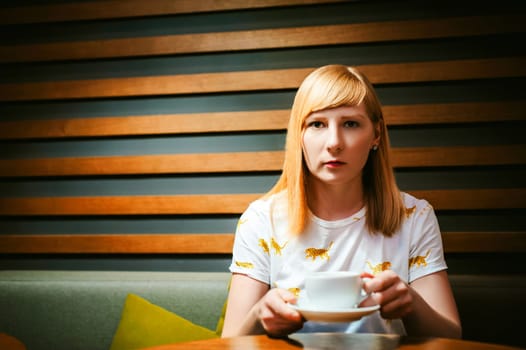Young blonde woman wearing white T-shirt with print, girl Drinks from a white porcelain cup holding it in his hands, sitting at a table in a cafe, background stylized Wood texture lit up with warm light