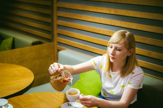 Young blonde woman wearing white T-shirt with print, girl pours a fruit drink from a glass jug, into a white cup, sitting at a table in a cafe, background stylized Wood texture lit up with warm light