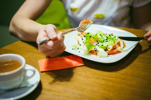 Young blonde woman wearing white T-shirt with print, girl Eats salad at lunch time, sitting at a table in a cafe, background stylized Wood texture lit up with warm light