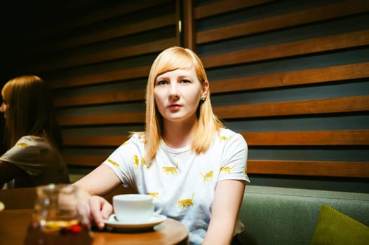 Young blonde woman wearing white T-shirt with print, girl Drinks from a white porcelain cup holding it in his hands, sitting at a table in a cafe, background stylized Wood texture lit up with warm light