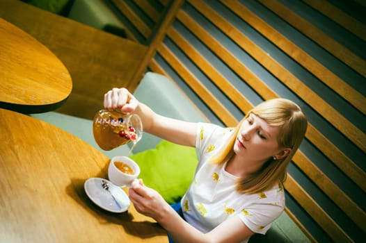 Young blonde woman wearing white T-shirt with print, girl pours a fruit drink from a glass jug, into a white cup, sitting at a table in a cafe, background stylized Wood texture lit up with warm light