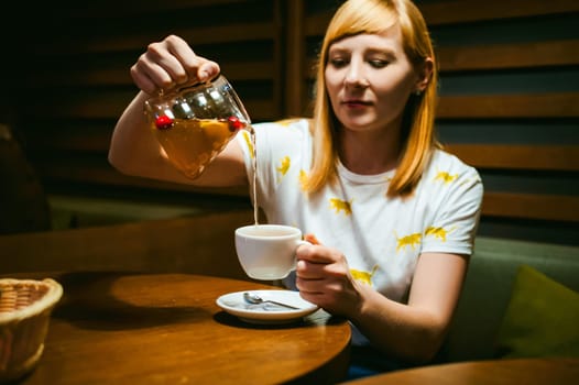Young blonde woman wearing white T-shirt with print, girl pours a fruit drink from a glass jug, into a white cup, sitting at a table in a cafe, background stylized Wood texture lit up with warm light