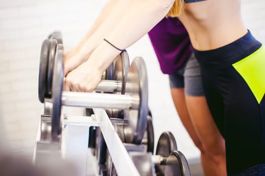 girl in a bright white gym. Two young athletic women near sports equipment are preparing for the traning with dumbbells, holding up in their hands. Healthy way of life, good spirits