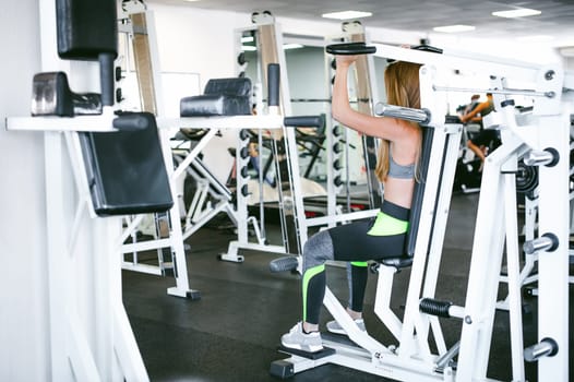 girl in a bright white gym. A blonde woman doing a strength exercise for a group of muscles on the hands of a simulator. Healthy way of life, good spirits