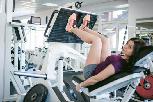girl in a bright white gym. A brunette woman doing a power exercise for a group of leg muscles on a simulator. Healthy way of life, good spirits