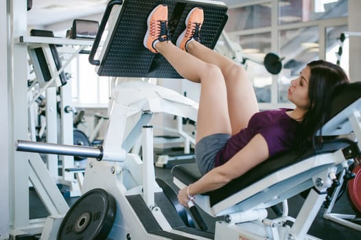girl in a bright white gym. A brunette woman doing a power exercise for a group of leg muscles on a simulator. Healthy way of life, good spirits
