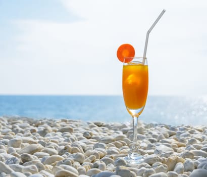 Orange juice with carrot slice in cocktail glass on white pebbly beach with seascape background, summer tropical holiday concept