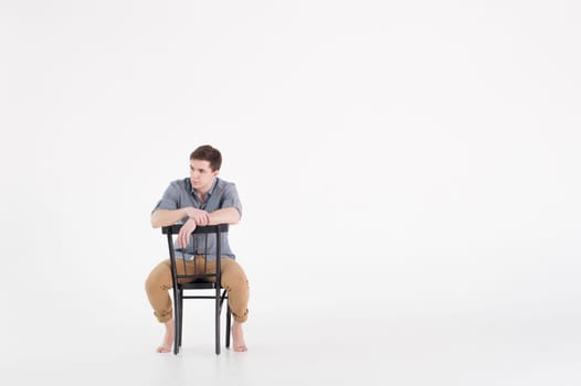 Man dressed in a shirt and brown trousers sits on a wooden vintage chair on a white background