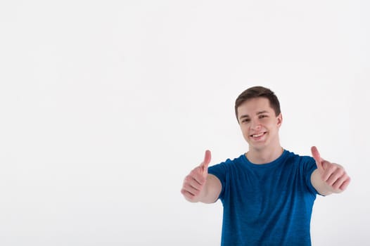 Young muscular handsome athletic man dressed in blue sport shirt standing on white background in studio, Shows thumbs up and smiles