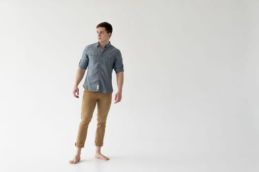 Young handsome man of strong athletic build, dressed in a shirt and brown trousers, Posing on white studio background